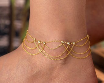 May include: A gold-colored anklet with multiple delicate chain layers and small, clear gemstones. The anklet is draped around the ankle, creating a layered effect. The jewelry is displayed on a foot, showcasing the design and fit.