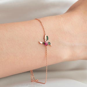 Könnte beinhalten: Ein zartes roségoldenes Armband mit einem Kolibri-Design. Der Kolibri ist mit grünen und rosa Edelsteinen verziert. Das Armband wird an einem Handgelenk gezeigt, um seine feinen Details und sein elegantes Design hervorzuheben.