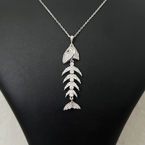 May include: A silver necklace with a fish bone pendant. The pendant is made of silver and is decorated with small, clear stones. The necklace is on a black display stand.