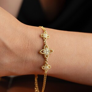 May include: Gold-toned bracelet featuring three clover-shaped charms. Each charm has a textured border and a central cluster of small, sparkling stones. The bracelet is worn on a wrist, with a black background.