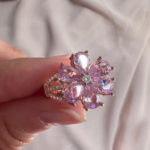 Puede incluir: Primer plano de un anillo con forma de flor. El anillo presenta una piedra central transparente rodeada de piedras rosas en forma de pera, creando los pétalos. La banda es dorada y está adornada con piedras más pequeñas y transparentes. El anillo es sostenido por una mano.