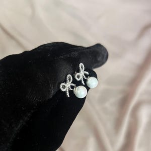 May include: A pair of silver-toned bow earrings with clear, sparkling accents and a dangling white pearl. The earrings are displayed against a black surface and a soft pink background.
