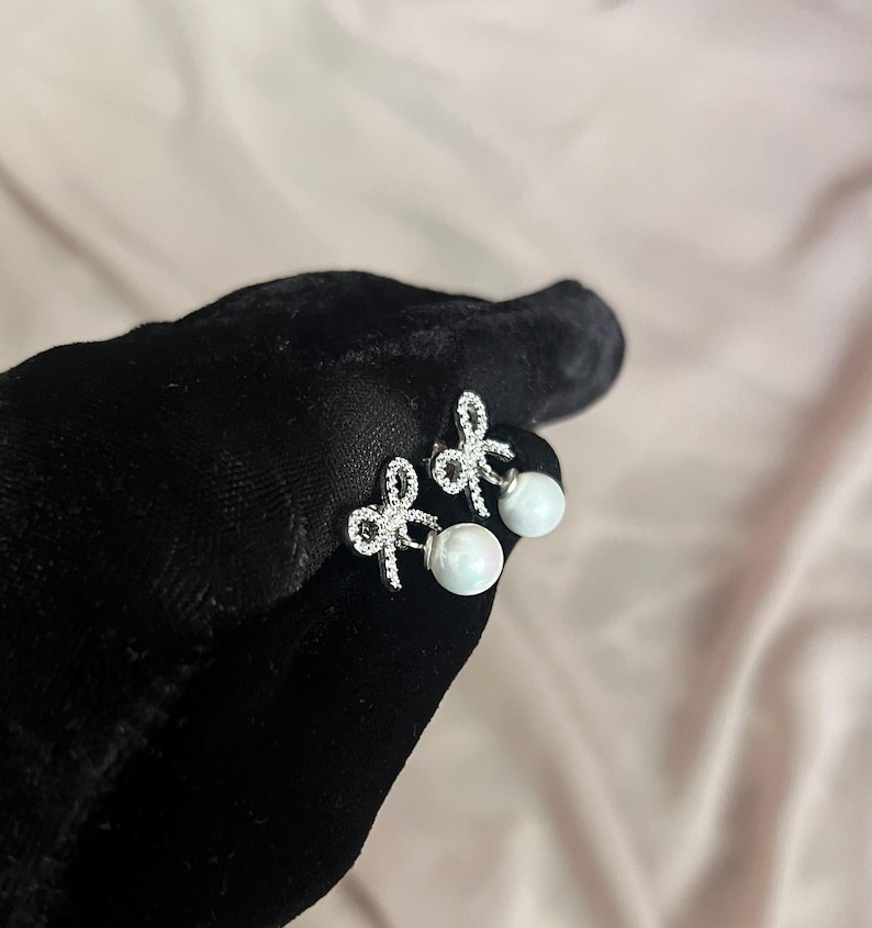 May include: A pair of silver-toned bow earrings with clear, sparkling accents and a dangling white pearl. The earrings are displayed against a black surface and a soft pink background.