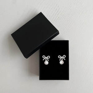 May include: A pair of bow-shaped earrings with a dangling pearl, presented in an open black box. The earrings feature a silver-colored frame with clear, sparkling accents. The box is on a white surface.