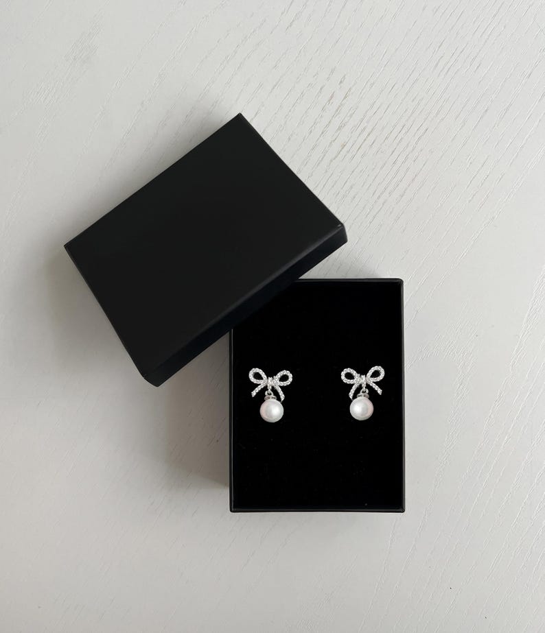 May include: A pair of bow-shaped earrings with a dangling pearl, presented in an open black box. The earrings feature a silver-colored frame with clear, sparkling accents. The box is on a white surface.