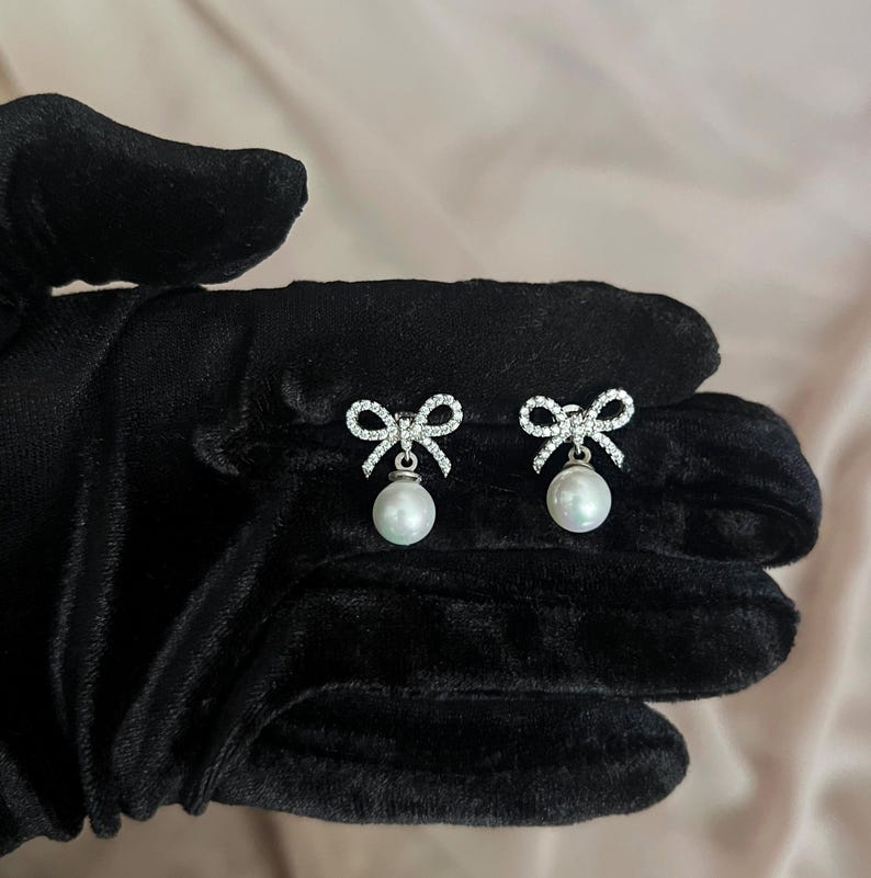 May include: A pair of silver-toned bow earrings with a dangling white pearl. The bows are encrusted with small, clear crystals. The earrings are displayed on a black surface.
