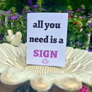 May include: A white rectangular greeting card featuring the black text "all you need is a" followed by the purple text "SIGN" and a small purple heart icon with wings and a halo, positioned upright on a white scalloped birdbath basin.