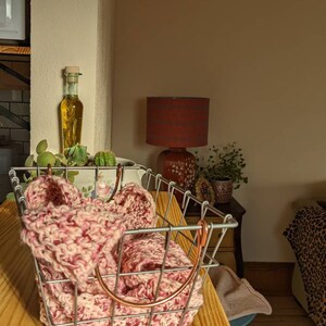 May include: A metal wire basket with a copper handle holds a pink and white speckled knitted item. A glass bottle with a cork stopper and a red lamp are visible in the background. The basket sits on a wooden surface.