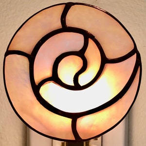 Könnte beinhalten: Ein weißes Nachtlicht mit einem Buntglas-Schirm, der ein Spiralmuster in Rosa- und Brauntönen zeigt. Die Lampe hat einen Ein-/Ausschalter.