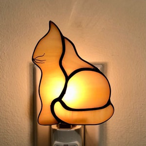 Può includere: Una luce notturna in vetro colorato a forma di gatto. Il gatto è seduto con la coda arrotolata attorno al suo corpo. Il vetro è di colore ambrato chiaro e la luce brilla attraverso il vetro.