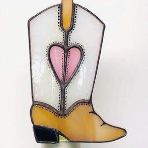 May include: A decorative night light shaped like a cowboy boot. The boot features a white upper with a pink heart design, a tan lower section, and a black heel. The night light is designed to plug into a standard electrical outlet.