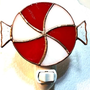 May include: A stained glass night light shaped like a round peppermint candy with alternating red and white segments. The glass pieces are joined by dark metal solder lines, and the light features small clear glass side accents and a white plug base.