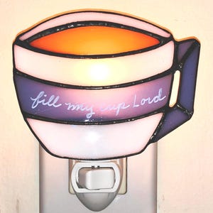 May include: A stained glass nightlight shaped like a coffee mug with the words "fill my cup Lord" written on it. The mug is white with purple and orange accents.