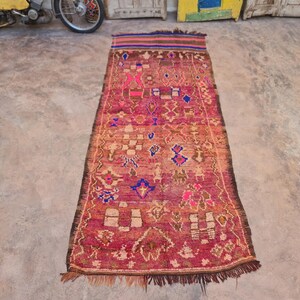 May include: A long, rectangular rug with a red background and intricate patterns in shades of blue, pink, and yellow. The rug has a fringe border and is made of woven wool.