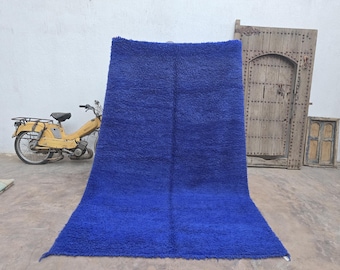Puede incluir: Una alfombra azul grande y rectangular con una superficie texturizada. La alfombra se exhibe en interiores, apoyada contra una pared. En el fondo se ve un ciclomotor amarillo, junto con una puerta y un marco de ventana de madera.