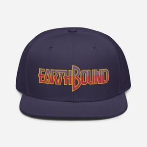 Puede incluir: Una gorra de béisbol azul marino con un logotipo "EARTHBOUND" bordado en amarillo, rojo y morado en la parte delantera.
