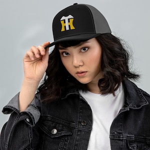 Könnte beinhalten: Eine schwarze und graue Trucker-Kappe mit einem weißen und goldenen gestickten Logo. Das Logo ist ein stilisiertes "HH" mit einem kleinen weißen Design über den Buchstaben.