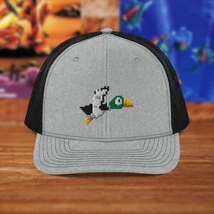 Puede incluir: Una gorra de camionero gris y negra con un pato pixelado bordado en la parte delantera. El pato está volando y tiene la cabeza verde y el pico amarillo.