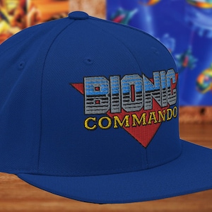 May include: A royal blue baseball cap with a red, white and blue embroidered logo that says "Bionic Commando".