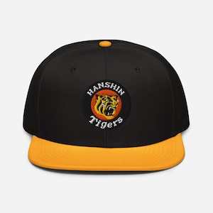 Könnte beinhalten: Schwarze und goldene Baseballkappe mit flachem Schirm. Auf der Kappe befindet sich ein kreisförmiger Aufnäher mit einem Tiger-Motiv und dem Schriftzug "HANSHIN Tigers" in Weiß. Die Krone ist schwarz, der Schirm goldfarben.