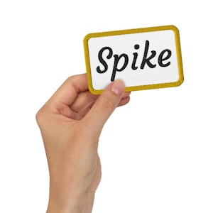 May include: A hand holds up a yellow-bordered white patch with the word "Spike" embroidered in black.