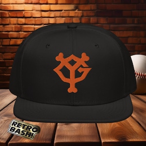 Puede incluir: Gorra de béisbol negra con un logotipo bordado en naranja de un "YG" estilizado dentro de un diseño en forma de hueso. La gorra tiene una visera curvada y una parte superior plana.