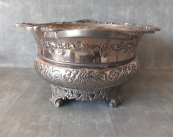 May include: A silver-colored, ornate serving bowl with a decorative floral and grape design. The bowl has a wide, scalloped rim and rests on three ornate feet. The bowl's surface reflects light, highlighting its intricate details.