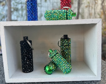 May include: Six small, jeweled, keychain containers in a white display box. The containers are in various colours, including red, blue, black, and green.