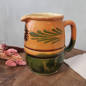 Könnte beinhalten: Eine antike Keramikkanne mit einem grünen und braunen Blumendesign. Die Kanne hat einen Henkel und eine breite Öffnung.