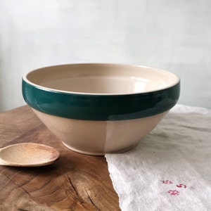 Può includere: Una grande ciotola per mescolare in ceramica color crema con un bordo verde scuro. La ciotola è appoggiata su una superficie di legno con un cucchiaio di legno accanto.