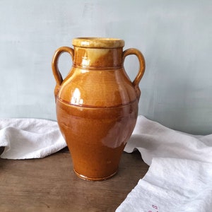 以下が含まれることがあります： 2つの取っ手のある大きな茶色の陶器のピッチャー。ピッチャーは釉薬仕上げで、わずかに丸みを帯びた形をしています。