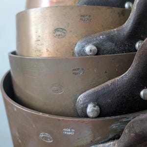 Puede incluir: Una pila de cacerolas de color cobre con asas marrones oscuras. Las cacerolas están encajadas unas dentro de otras, con las asas extendiéndose hacia afuera. La imagen muestra las palabras "MADE IN FRANCE" estampadas en el lateral de las cacerolas.