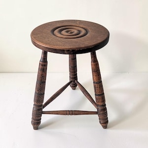 May include: A small, three-legged wooden stool with a dark brown finish. The circular seat features a carved concentric ring pattern in the center. The three legs are turned with decorative ridges and are connected by a triangular support frame.