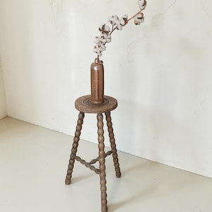 May include: A tall, wooden stool with a round seat and three legs featuring a decorative bobbin-turned design. A brown ceramic bottle sits on the center of the seat, holding a single dried branch with white cotton bolls extending upward.