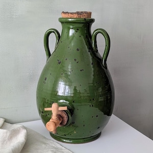 May include: A green ceramic water jug with a cork stopper and a wooden spigot. The jug has two handles and a speckled finish. The jug is sitting on a white surface.