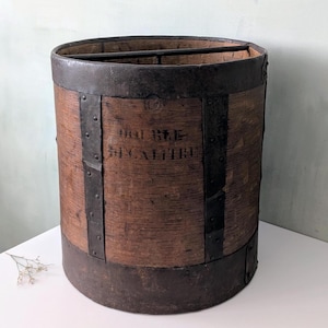 Peut inclure: Une mesure à grain cylindrique en bois avec des bandes et des rivets en métal sombre. Le bois présente une texture brune patinée et l'inscription noire DOUBLE DECALITRE. Une fine barre métallique traverse l'ouverture supérieure du récipient.