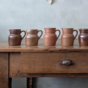 Peut inclure: Une collection de pichets en céramique faits à la main, dans diverses nuances de brun, du beige clair au chocolat foncé. Les pichets sont disposés sur une table en bois rustique, mettant en valeur leurs formes et tailles uniques. Un tissu en lin est suspendu en arrière-plan.
