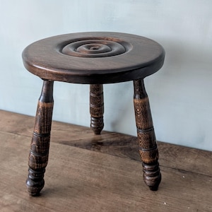 May include: A small, dark brown wooden stool with a round seat featuring a spiral design. The stool has three turned legs. The wood grain is visible, giving it a rustic appearance. The stool is likely vintage or antique.