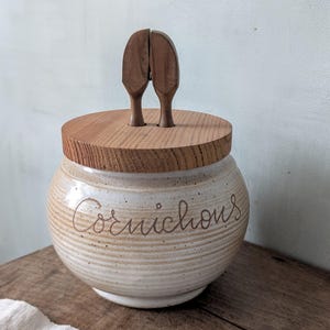 Op de afbeelding: Een beige keramische pot met een houten deksel en handvat. Op de pot staat het woord "Cornichons". Het deksel is rond en gemaakt van lichtbruin hout. Het handvat bestaat uit twee houten stukken die met elkaar verbonden zijn.