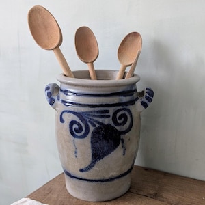 May include: A beige stoneware utensil holder with a blue floral design and two small handles, containing three wooden spoons. The holder sits on a wooden surface.
