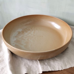 Może przedstawiać: Płytka, okrągła ceramiczna miska o gładkim, matowym wykończeniu w ciepłym, piaskowo-beżowym odcieniu. Wewnętrzna powierzchnia posiada subtelne, jaśniejsze kremowe wariacje szkliwa, tworzące miękki, chmurowy wzór na dnie naczynia.