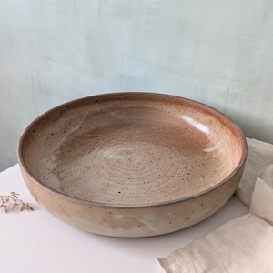 Peut inclure: Un grand bol en céramique fait à la main, de forme large et peu profonde. Le bol présente une palette de couleurs terreuses et mouchetées, avec des nuances de brun et de beige. L'intérieur est lisse et clair, tandis que le bord et l'extérieur sont plus foncés.