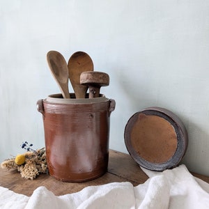 Könnte beinhalten: Ein brauner Keramiktopf mit Deckel, gefüllt mit Holzlöffeln und einem Holzgriff. Der Topf steht auf einem Holztisch mit einem weißen Tuch darüber.