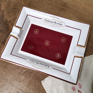 Op de afbeelding: Witte asbak van keramiek met een gouden rand en een rood interieur. Het interieur heeft de tekst "Champagne Laurent Perrier" en een herhalend logo van de letter "LP" in een cirkel.