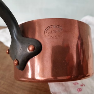 May include: A copper saucepan with a dark metal handle attached by three copper rivets. The side of the pan features a stamped oval mark that reads GRANDS MAGASINS DU LOUVRE PARIS. The surface of the copper is smooth and reflective.