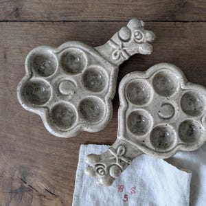 Pode incluir: Dois pratos de servir de cerâmica em forma de camelo com seis reentrâncias individuais para servir ovos ou outros petiscos pequenos. Os pratos são de cor cinza claro salpicado.