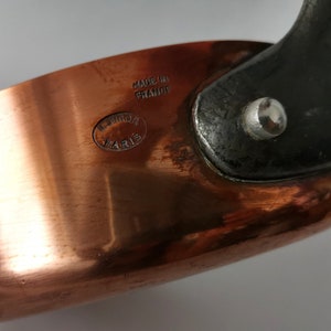 Peut inclure: Une casserole en cuivre avec une poignée noire et un rivet argenté. Le fond de la casserole est estampillé "Made in France" et "Cuivron Paris".