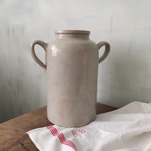 Könnte beinhalten: Eine große, graue Keramikvase mit zwei Henkeln. Die Vase hat eine leicht strukturierte Oberfläche und steht auf einem weißen Leinenstoff mit roten Streifen.