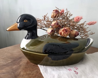 May include: A ceramic duck-shaped container with a black head, a yellow beak, and a green and black body. The container is decorated with dried flowers. The duck is sitting on a wooden surface.