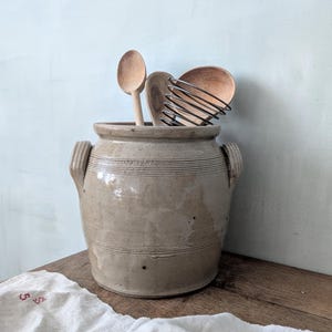 Puede incluir: Un tarro de cerámica beige con líneas horizontales y dos asas laterales, lleno de cucharas de madera y un batidor. El tarro está sobre una superficie de madera, con un paño blanco en primer plano. El fondo es una pared azul claro.